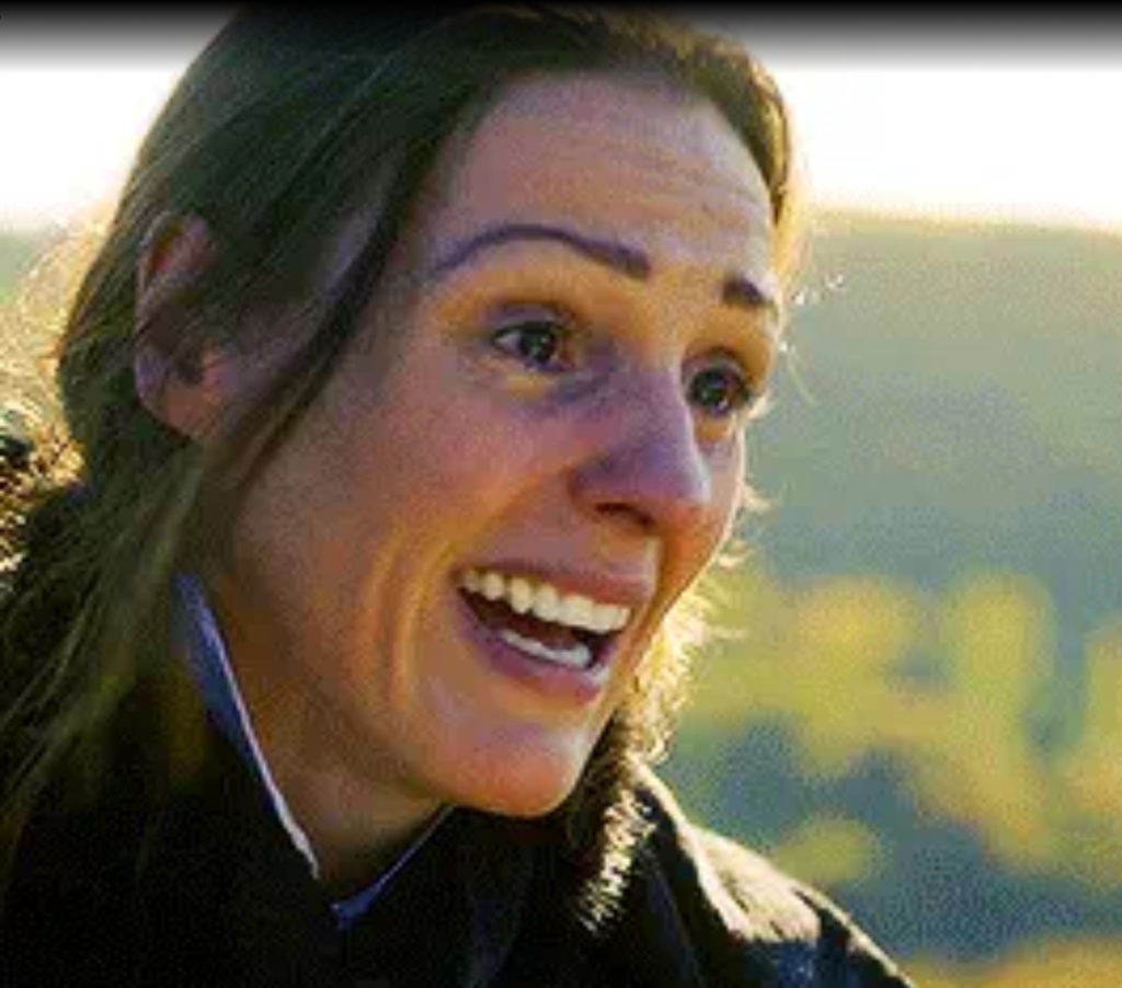 Suranne Jones, playing Anne Lister, with a big, expectant smile on her face. Bright green hills blurred in the background. 